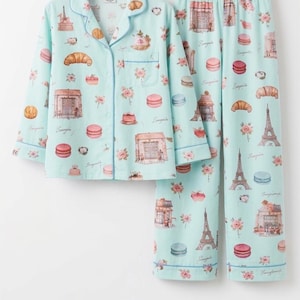 May include: A light blue pajama set with a button-down top and trousers. The set features a repeating pattern of Parisian landmarks, croissants, macarons, and floral accents. The pajamas are trimmed with contrasting blue piping. The set is made of a soft material.