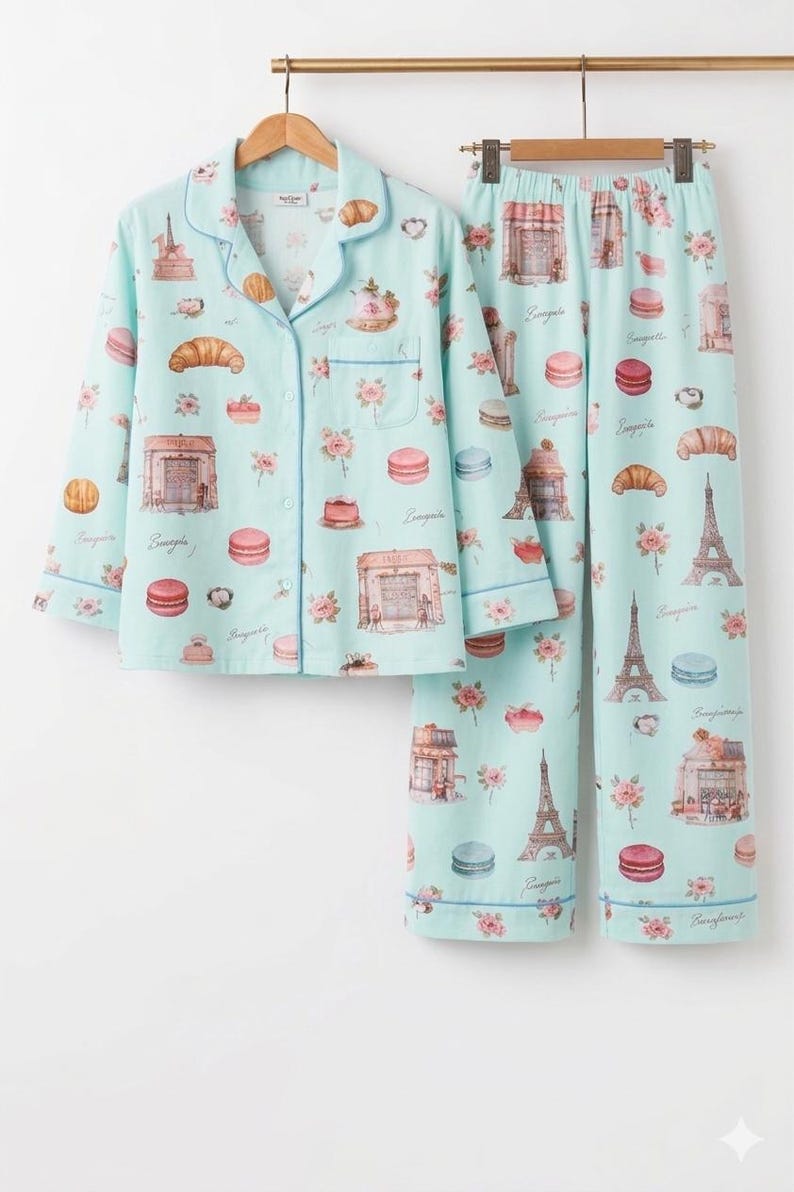 May include: A light blue pajama set with a button-down top and trousers. The set features a repeating pattern of Parisian landmarks, croissants, macarons, and floral accents. The pajamas are trimmed with contrasting blue piping. The set is made of a soft material.