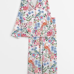 May include: A white pajama set featuring a long-sleeved button-down shirt and matching pants. The fabric is decorated with a colorful pattern of birds, flowers, and mahjong tiles with Chinese characters, accented by thin pink piping trim.