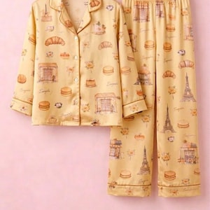 May include: Yellow pyjama set featuring a pattern of croissants, macarons, and the Eiffel Tower. The long-sleeved top has a button-down front and a notched collar. The trousers have an elasticated waistband. The set is hanging on a wooden hanger.