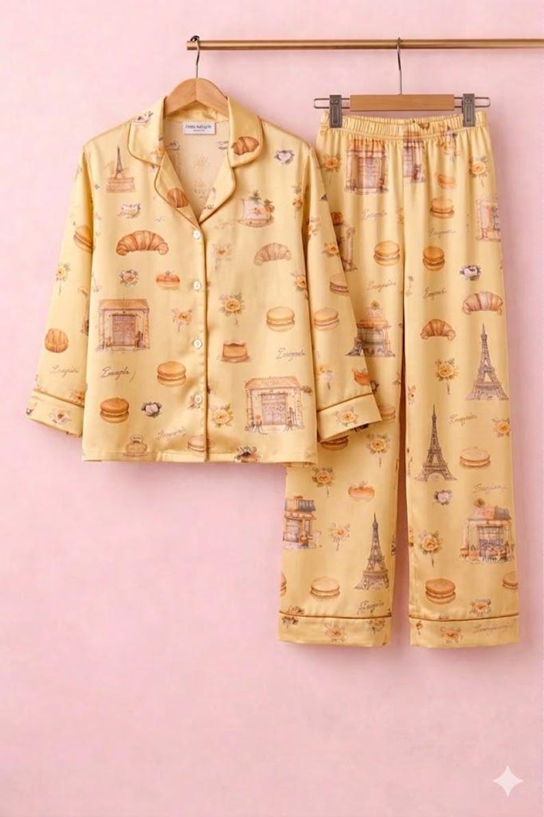 May include: Yellow pyjama set featuring a pattern of croissants, macarons, and the Eiffel Tower. The long-sleeved top has a button-down front and a notched collar. The trousers have an elasticated waistband. The set is hanging on a wooden hanger.