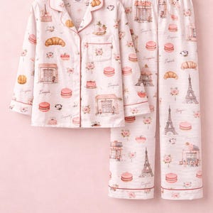 May include: Light pink pyjama set with a button-up shirt and trousers. The set is patterned with Parisian imagery, including the Eiffel Tower, croissants, and macarons. The edges are trimmed with a darker pink. Measurements are in cm.