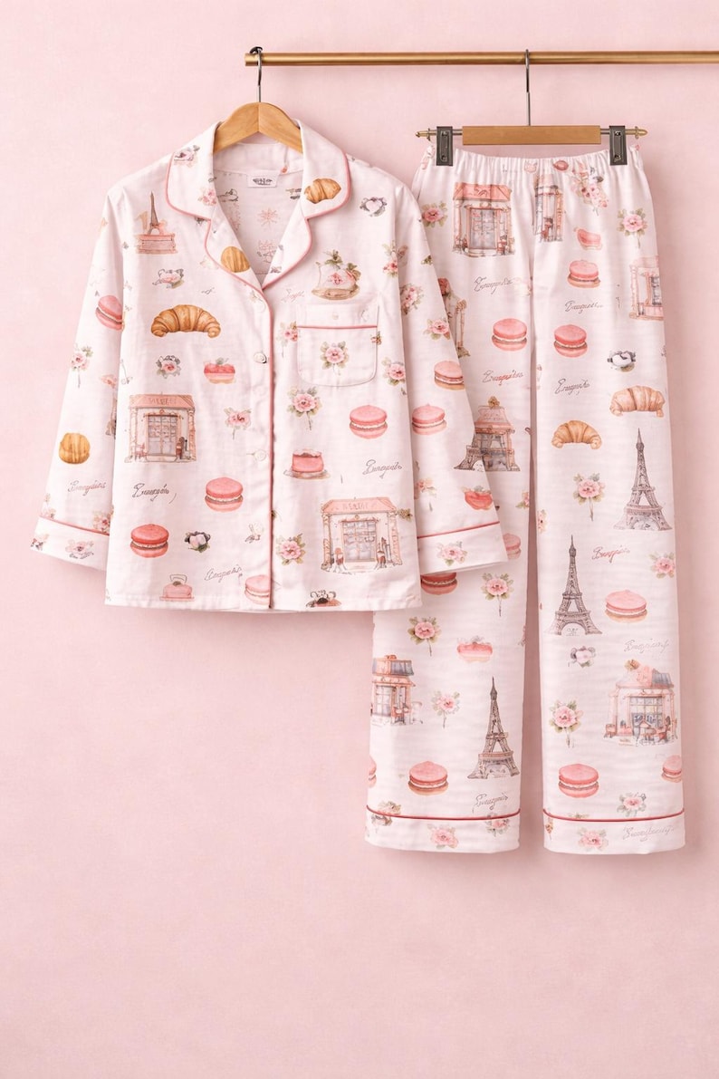 May include: Light pink pyjama set with a button-up shirt and trousers. The set is patterned with Parisian imagery, including the Eiffel Tower, croissants, and macarons. The edges are trimmed with a darker pink. Measurements are in cm.