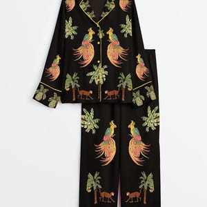 May include: A black pajama set featuring a long-sleeved button-down shirt with a notched collar and matching wide-leg pants. The set is decorated with an embroidered tropical pattern of colorful parrots, palm trees, monkeys, and pineapples.