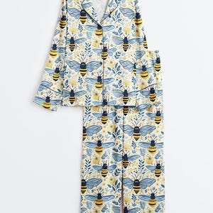 May include: A two-piece pajama set featuring a long-sleeved button-down shirt with a notched collar and matching long pants. The fabric is white and decorated with a repeating pattern of yellow and black bees, blue wings, and small yellow flowers.