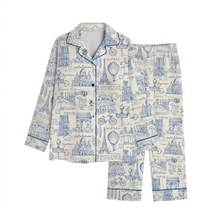 May include: Cream-coloured two-piece pyjama set with a blue Parisian-themed print. The long-sleeved top has a button-down front and a notched collar. The matching trousers have an elasticated waistband. The print includes the Eiffel Tower, Arc de Triomphe, and text.