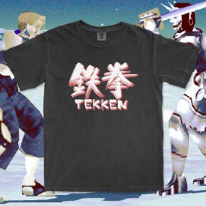 Puede incluir: Una camiseta de manga corta de color gris carbón que presenta los caracteres kanji japoneses de Tekken en blanco con un sutil efecto de sombra roja, situados sobre la palabra TEKKEN en letras mayúsculas blancas con una sombra roja a juego.