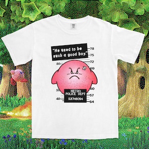 May include: A white short-sleeve t-shirt featuring a graphic of a pink, round, angry character with a bandage on its head. The design includes a mugshot height chart, the text "He used to be such a good boy," and "METRO POLICE DEPT K8746094."