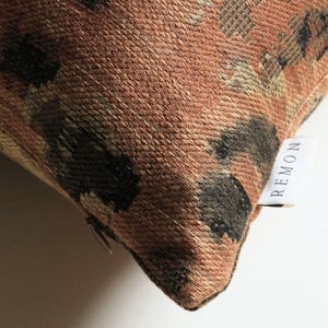 May include: A square throw pillow featuring a textured, woven fabric with a pattern of abstract dark brown and tan shapes on a rust-colored background. A small white fabric tag with the black text REMON is sewn into the side seam near a zipper.