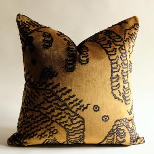 May include: A square throw pillow featuring a golden-brown velvet fabric base with a dark charcoal black embroidered pattern. The embroidery consists of dense, textured, repeating geometric lines and circular motifs across the front surface.