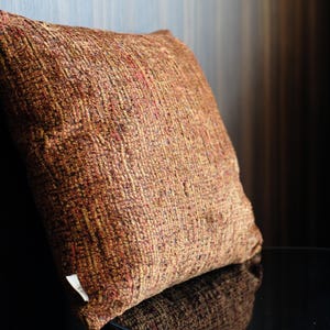 Puede incluir: Un cojín cuadrado con una tela texturizada multicolor en tonos óxido, marrón y dorado. El material tejido tiene un patrón de superficie visible, grueso e irregular. Una pequeña etiqueta de tela blanca está unida a la esquina inferior izquierda.
