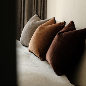 May include: Three square throw cushions arranged in a row, featuring a textured fabric in shades of light grey, medium brown, and dark brown. Each cushion is finished with a thin, dark piping along the edges, creating a defined border.