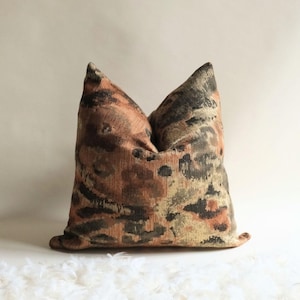 Square decorative pillow with textured abstract pattern in warm rust and olive tones, styled against a neutral background