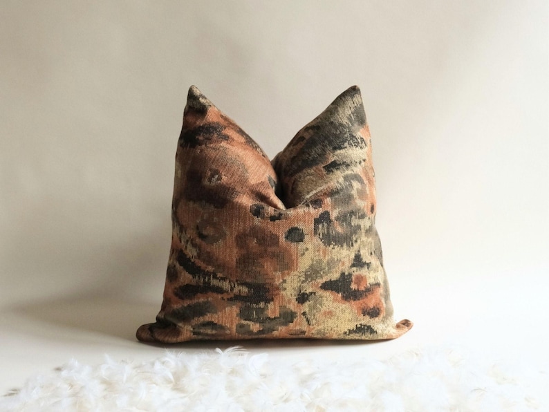 Square decorative pillow with textured abstract pattern in warm rust and olive tones, styled against a neutral background