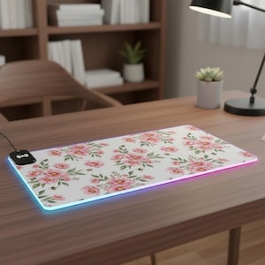May include: A rectangular mouse mat with a floral pattern of pink roses and green leaves on a white base. The mat has a colour-changing edge. A black wireless charging receiver is connected to the mat.