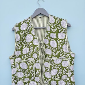 May include: A sleeveless quilted waistcoat featuring a vibrant olive green and white floral pattern with large chrysanthemum-like blooms. The garment includes two front patch pockets, a tie-front closure, and striped trim along the edges and interior.
