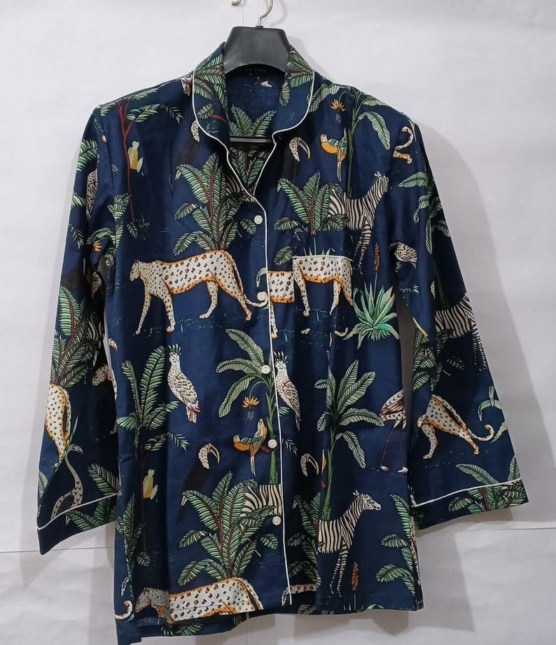 Puede incluir: Top de pijama azul oscuro con estampado de animales de la jungla con leopardos, cebras y loros. La parte superior de manga larga tiene ribetes blancos a lo largo del cuello, los pu&ntilde;os y la tapeta de los botones. La tela tiene un estampado de palmeras y otro follaje tropical.