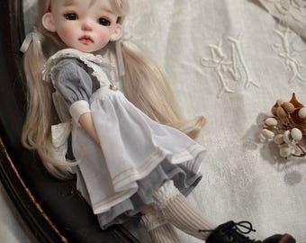 May include: A ball-jointed doll with long blonde hair styled in pigtails, wearing a grey dress with a white pinafore, lace collar, beige ribbed socks, and black lace-up boots. The doll has large dark eyes and a neutral expression on its face.