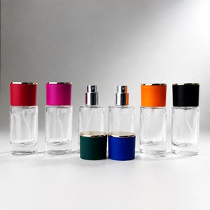 Pode incluir: Seis frascos de vidro transparente com atomizador, corpos cilíndricos e bicos metálicos. Quatro frascos possuem tampas de cores sólidas em vermelho, magenta, laranja e preto, enquanto duas tampas verde-escuras e azuis estão à frente.