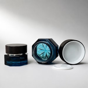May include: Three octagonal cosmetic jars made of deep blue glass. One jar is closed with a dark wooden lid, one is open showing a faceted interior, and one is shown with its black screw-top lid removed, revealing a white plastic inner liner.
