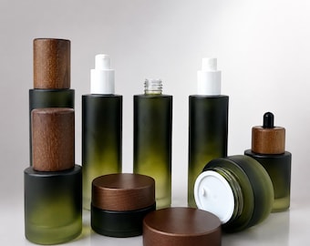Op de afbeelding: Een collectie cosmetische verpakkingen van mat donkergroen glas, bestaande uit cilindrische flessen met witte pompdoseerders, een fles met schroefdop en potjes met donkerbruine houten deksels, waarvan er één op zijn zijkant ligt.