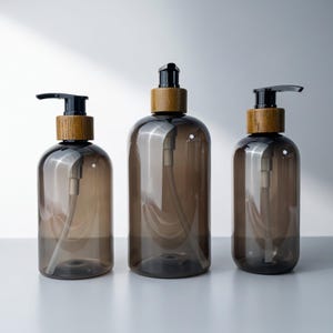 May include: Three translucent dark brown plastic bottles of varying sizes, each featuring a black pump dispenser mechanism and a decorative wooden collar. The bottles have a cylindrical shape and are arranged in a row on a flat surface.