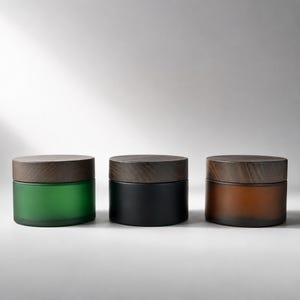 May include: Three cylindrical glass jars with dark brown wooden lids. The jars feature a frosted finish in green, black, and amber colors. Each container has a wide, flat base and a matching circular wooden cap that sits flush on the top.