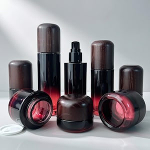 May include: A collection of cosmetic containers featuring dark red to black gradient glass bodies and dark brown wood-grain textured caps. The set includes tall pump bottles, cylindrical bottles with lids, and wide-mouth jars with matching lids.