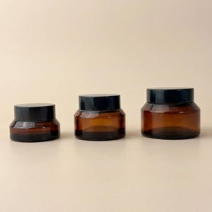 Op de afbeelding: Drie amberkleurige glazen potten met zwarte deksels staan op een rij. De potten variëren in grootte, met de kleinste links en de grootste rechts. De potten zijn leeg en staan op een neutraal gekleurd oppervlak.