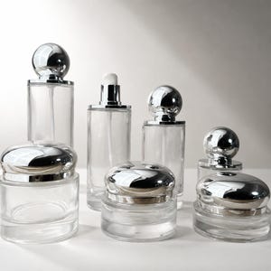 May include: A collection of clear glass cosmetic containers featuring cylindrical bottles with silver spherical caps, pump dispensers, and low-profile jars with rounded silver lids. The set includes various sizes of glass vessels for skincare.