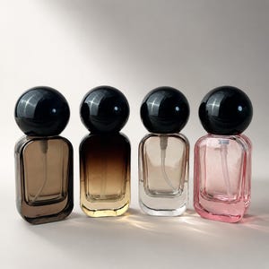 May include: Four rectangular glass perfume bottles with rounded corners and black spherical caps. Each bottle features a distinct color gradient: dark brown, amber, clear, and light pink. A clear plastic spray tube is visible inside each bottle.