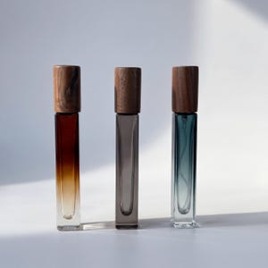 Puede incluir: Tres frascos de perfume rectangulares de vidrio con tapas de madera. Los frascos contienen líquidos ámbar, gris y verde azulado. Las tapas de madera marrón oscuro están sobre los frascos. Dispuestos en fila sobre un fondo blanco.