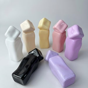 May include: A collection of seven glossy, irregular-shaped bottles with matching cube-shaped caps. The bottles are arranged in a group and come in a variety of solid colors including white, cream, yellow, pink, lavender, and black.