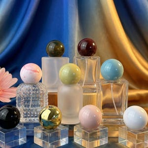 Puede incluir: Una variedad de frascos de perfume con tapones esféricos decorativos en varios colores, incluyendo verde, rojo, azul, rosa y dorado. Los frascos se exhiben sobre bloques acrílicos transparentes contra un fondo de tela azul y dorada.