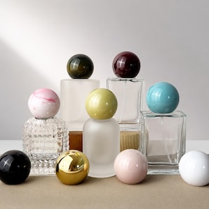 Può includere: Una collezione di flaconi di profumo in vetro con tappi sferici in vari colori e finiture, tra cui vetro trasparente, satinato e testurizzato. I tappi sferici presentano superfici solide, marmorizzate e metalliche in nero, rosa, oro, verde e blu.