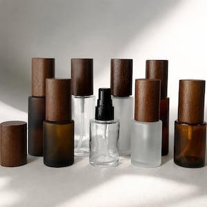 Puede incluir: Una colección de frascos pulverizadores cilíndricos de vidrio con tapas de madera marrón oscuro. Los frascos varían en acabado, incluyendo vidrio transparente, esmerilado y ámbar. Un frasco central transparente tiene un mecanismo de bomba negro.