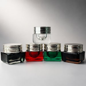 May include: Five square glass cosmetic jars with silver-toned lids arranged in a pyramid formation. The jars are colored in clear, black, red, green, and amber, each featuring a thick base and a multi-ridged metallic cap design.