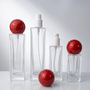 Puede incluir: Cuatro frascos rectangulares de vidrio transparente con dispensadores de bomba blancos. Dos frascos tienen grandes tapas esféricas de color rojo brillante, mientras que una esfera roja se encuentra sola en primer plano. Los frascos varían en altura.