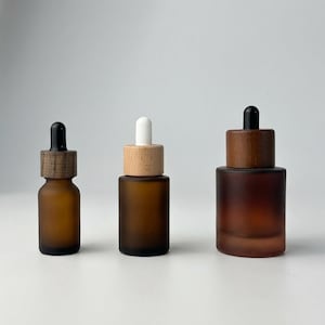 Può includere: Tre flaconi contagocce in vetro marrone con tappi in legno e plastica nera o bianca. I flaconi sono di diverse dimensioni, con finitura satinata. I tappi hanno un aspetto naturale venato del legno.