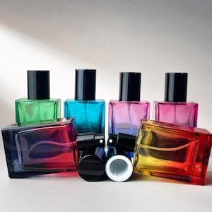 May include: An assortment of colorful glass perfume bottles with black spray tops. The bottles are in various shades, including green, blue, pink, and yellow, with a gradient effect. The bottles are arranged in a group.