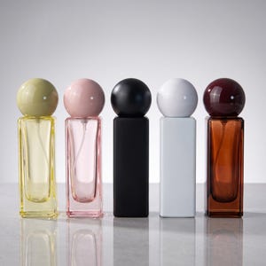 May include: Five rectangular glass perfume bottles arranged in a row. Each bottle features a spherical cap. The bottles are colored yellow, pink, black, white, and brown, with matching spherical caps. The black and white bottles are opaque.