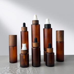 May include: A collection of eight amber glass bottles of varying heights and widths, each topped with a wooden cap. Some bottles feature white spray nozzles, while others are fitted with black dropper tops, creating a set of cosmetic containers.