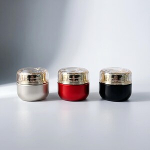 May include: Three small cosmetic jars arranged in a row. The first jar is matte silver, the second is glossy red, and the third is glossy black. Each jar features a clear, faceted, diamond-like lid with a gold-toned metallic band at the base.