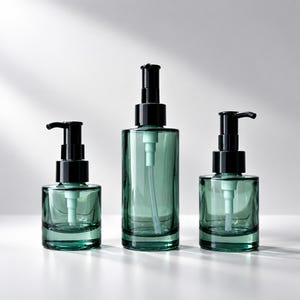 May include: Three cylindrical glass pump bottles in a deep emerald green hue. Each bottle features a black plastic pump mechanism with a light green internal tube. The set includes one tall central bottle and two shorter bottles of equal size.