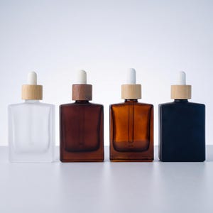 May include: Four square glass bottles with wooden and white dropper tops. The bottles are in different colors: clear, brown, amber, and black. The bottles are arranged in a row.