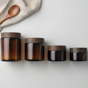 以下が含まれることがあります： さまざまなサイズの木製蓋が付いた4つの琥珀色のガラス瓶が並んでいます。瓶は円筒形で、濃い茶色の色合いです。木のスプーンとベージュの布が背景にあります。