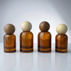 Puede incluir: Cuatro frascos pulverizadores de vidrio ámbar dispuestos en fila, cada uno rematado con una tapa esférica de madera. Los frascos tienen forma cilíndrica y base de vidrio transparente, mientras que las esferas de madera varían de tono claro a oscuro.