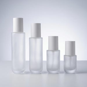 Könnte beinhalten: Vier Flaschen aus mattiertem Glas mit weißen Kappen unterschiedlicher Höhe. Die Flaschen sind in einer Reihe angeordnet, wobei die höchste Flasche links und die kürzeste rechts steht. Die Flaschen sind leer und scheinen für Kosmetikprodukte bestimmt zu sein.