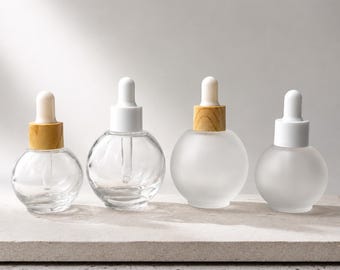 May include: Four round glass dropper bottles arranged in a row. Two bottles are made of clear glass, one with a light wood collar and one with a white collar. Two bottles are made of frosted glass, one with a light wood collar and one with a white collar.