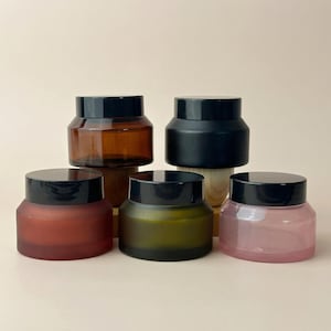 Pode incluir: Cinco potes cosméticos com tampas pretas. Os potes são de várias cores, incluindo âmbar, preto, vermelho, verde e rosa. Os potes são dispostos sobre um fundo neutro, mostrando suas diferentes tonalidades e texturas.